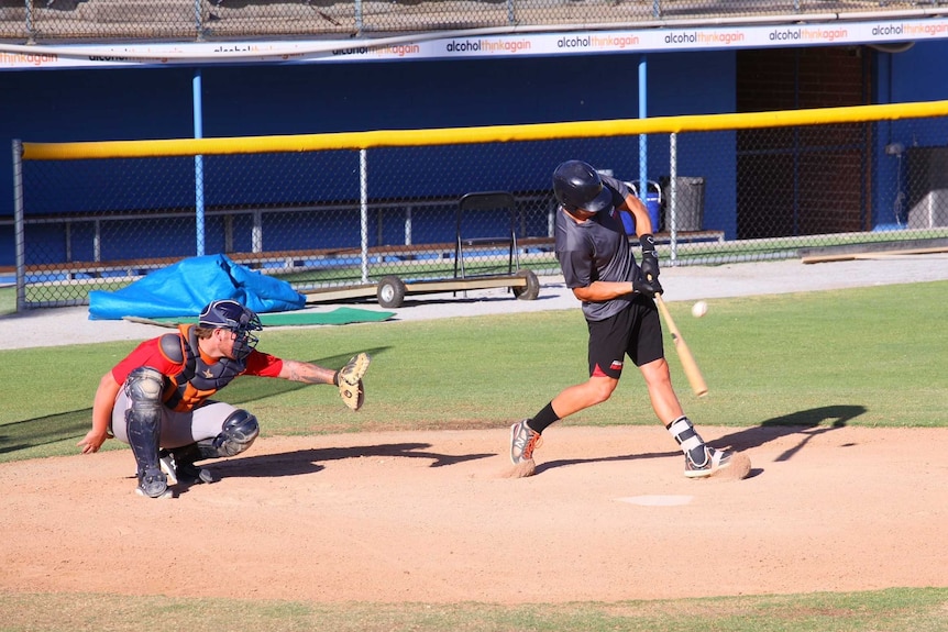 A side-on shot of a batter at the plate on a baseball field swinging at a ball with a catcher poised behind him.