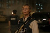 A woman stares ahead in a rioting city, wearing a blue press jacket sayign press. She has a camera around her.