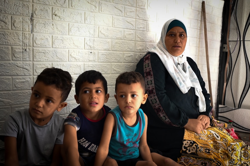 A woman wearing white headwrap and patterned skirt sits against a white wall next to three children
