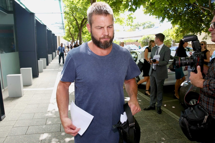 A mid shot of Ben Cousins walking on a footpath wearing a blue t-shirt with a cameraman beside him.