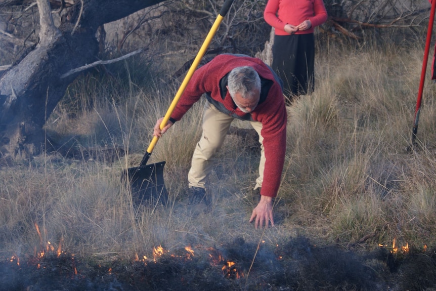 A man holding shovel inspects small flames burning in a line through grassland.