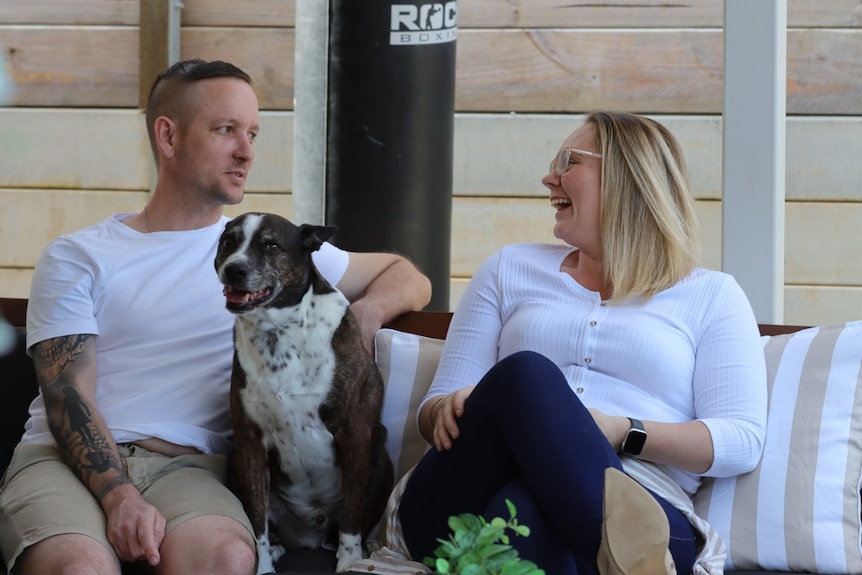 A man and woman sit together on an outdoor sofa, with their dog sitting between them.