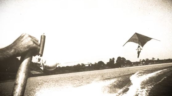 A black and white image of a man in a hang-glider soaring over a river behind a boat.