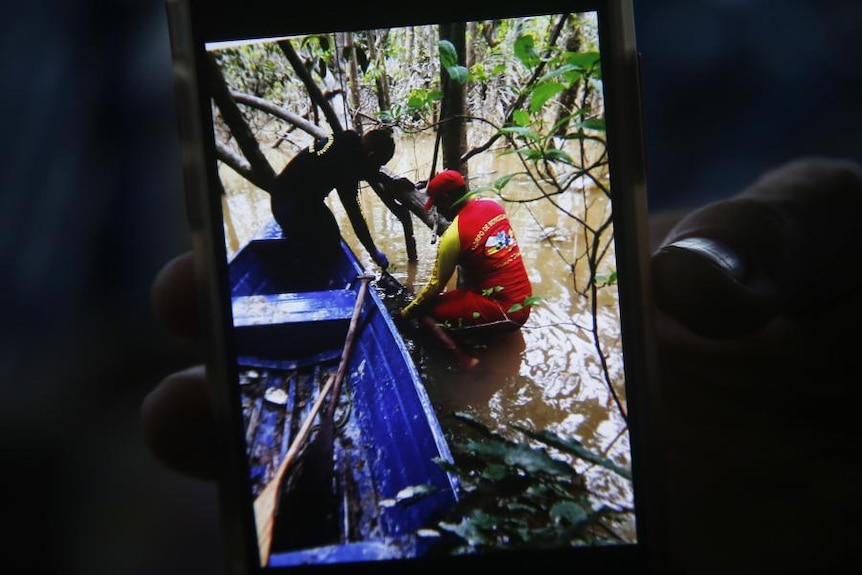 A person holds up a cell phone with a picture showing two rescue workers on a boat in the Amazon rainforest.