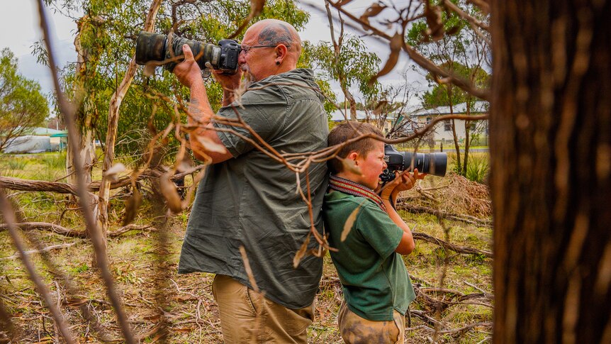 A man and young boy stand back-to-back in the bush, each holding a camera with a long lens