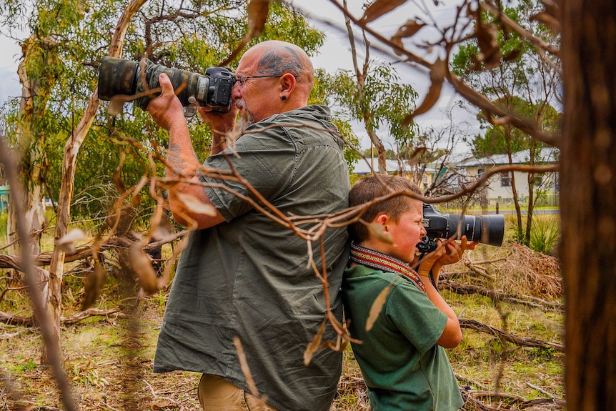 A man and young boy stand back to back holding large cameras in the bush.