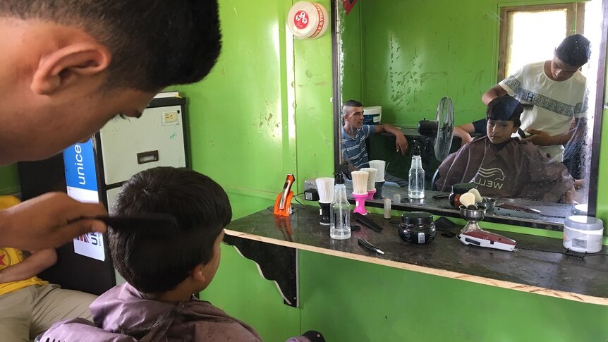 16-year-old Arabi cuts a young boy's hair while his customer looks on in a mirror.