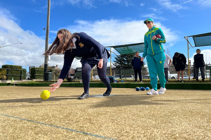 A student rolls a yellow bowl on a bowling green with Ellen Ryan watching on from behind.