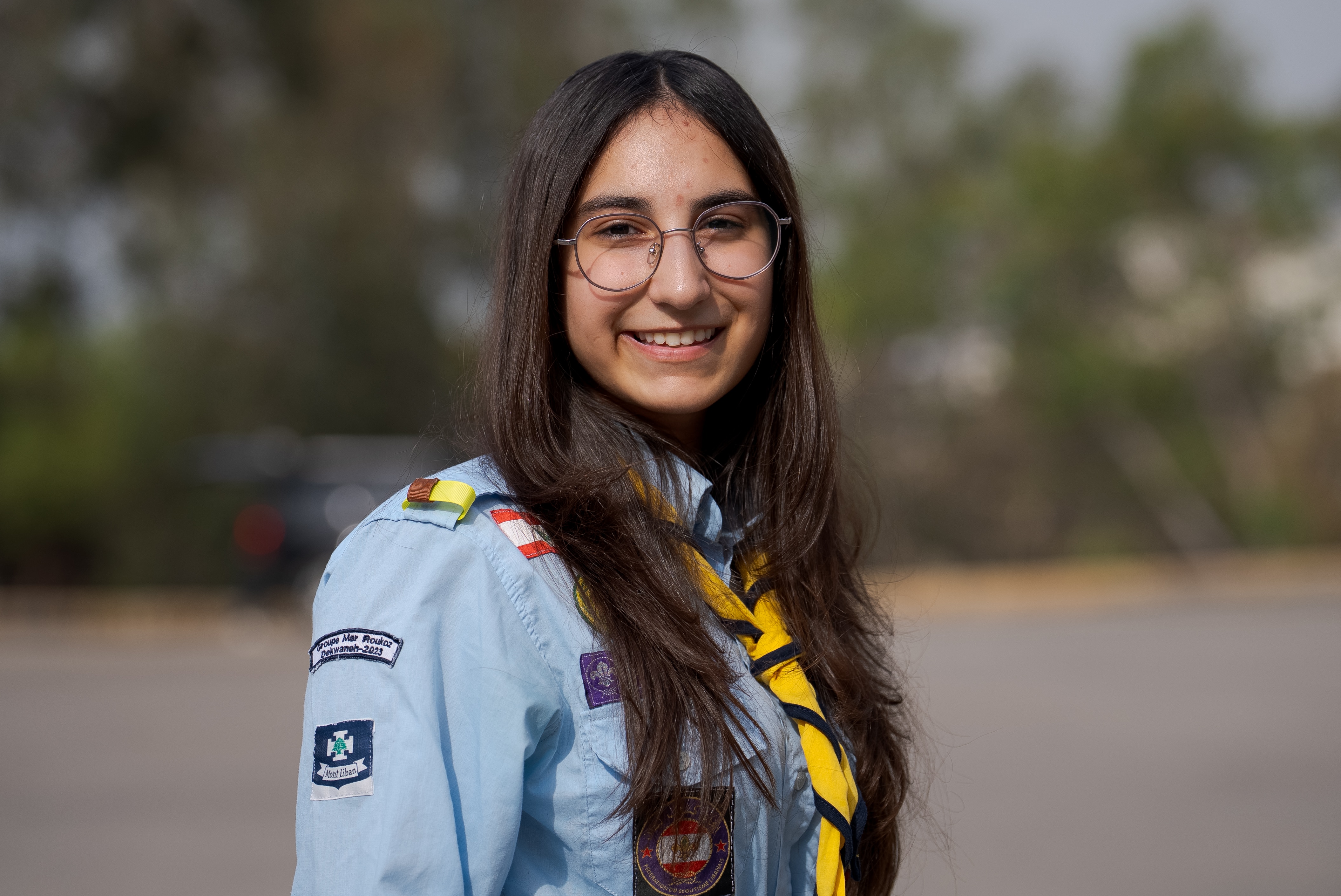 A young girl wearing glasses wears a scouts uniform with several patches and a scarf