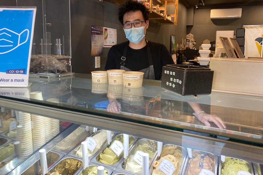 A man wearing a mask stands behind an array of gelato in a glass case.
