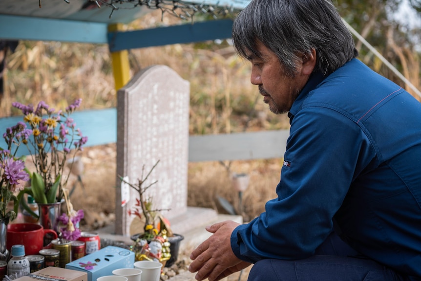 Man kneeling at a grave with flowers on it.