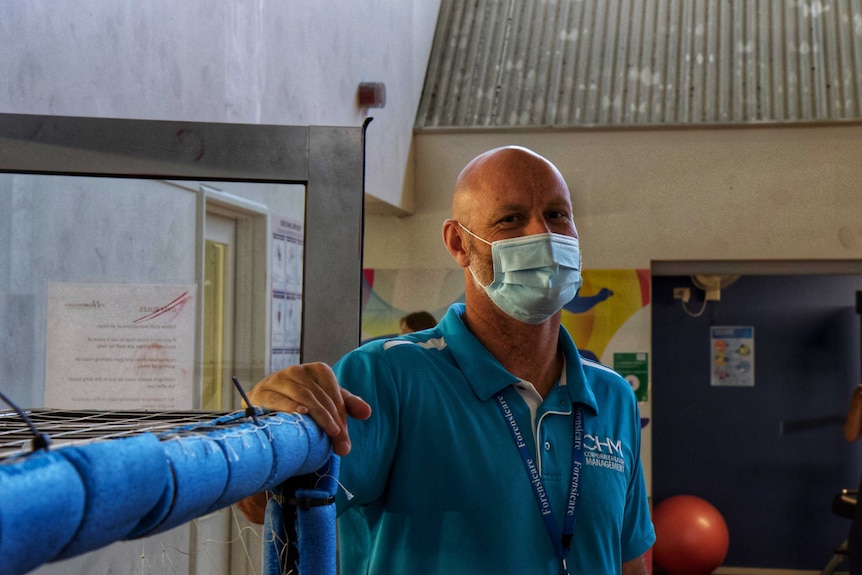 A man wearing a mask looks at the camera standing in a room. Behind him is a gym ball, next to him appears to be a soccer goal.