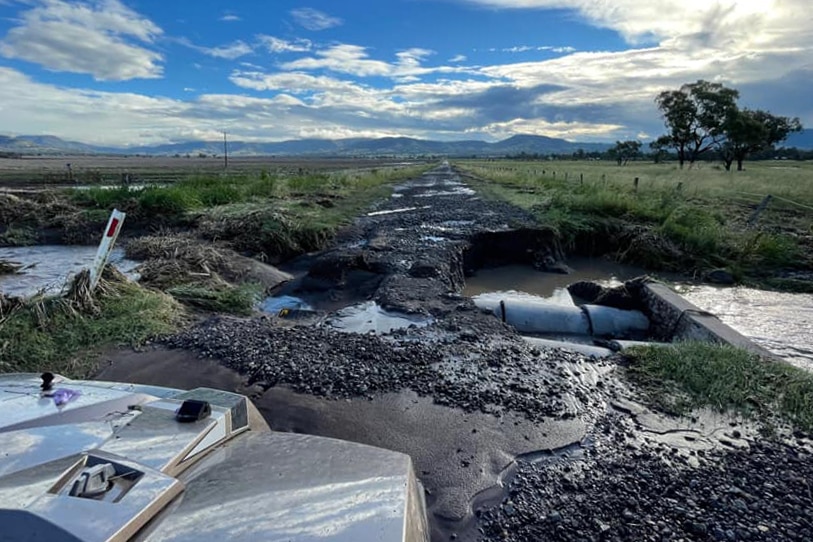 Badly cracked and damaged rural road with large potholes filled with water under a blue sky with surrounding green grass paddock