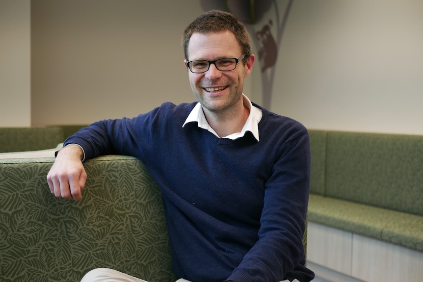 A man wearing spectacles sits down inside smiling for a photo with his legs crossed.