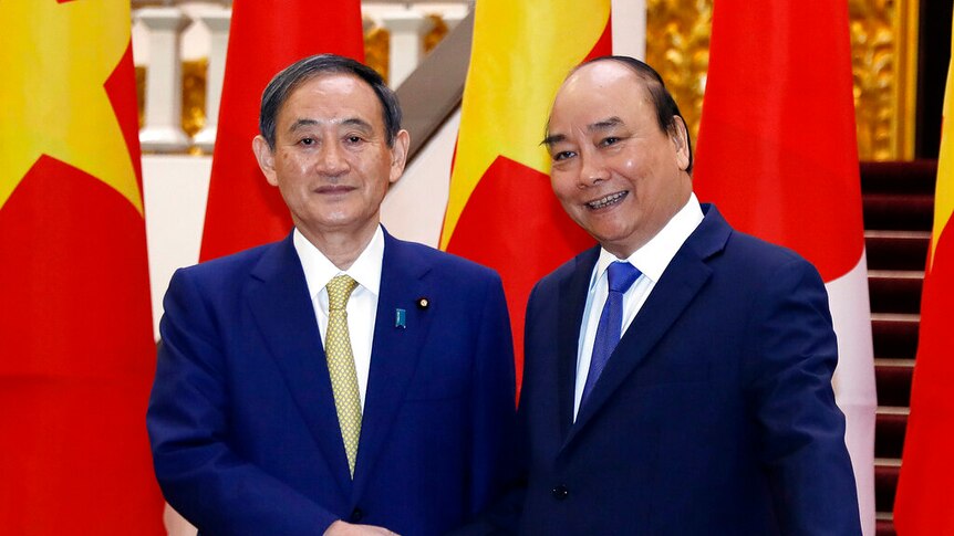 Yoshihide Suga and Nguyen Xuan Phuc shake hands in navy blue suits in front of large Vietnamese and Japanese flags.