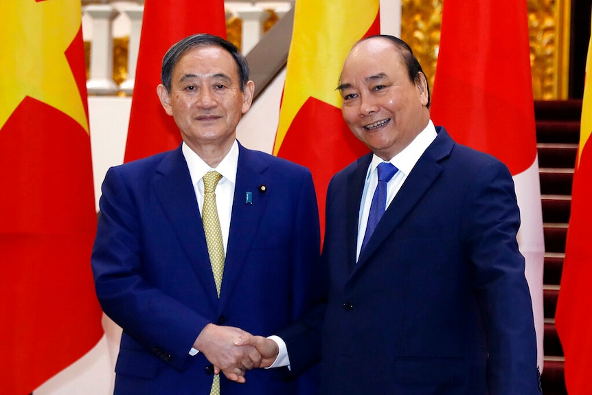 Yoshihide Suga and Nguyen Xuan Phuc shake hands in navy blue suits in front of large Vietnamese and Japanese flags.