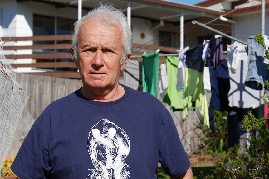 An elderly man with receding grey hair man in a dark blue t-shirt stands in front of a clothes line.