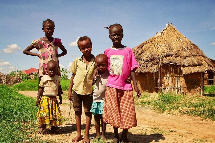 Five children stand on a dirt road in front of a grass and mud hut.