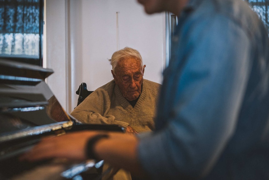An elderly man in a tan cardigan listens while a person in a blue shirt plays piano in the foreground.