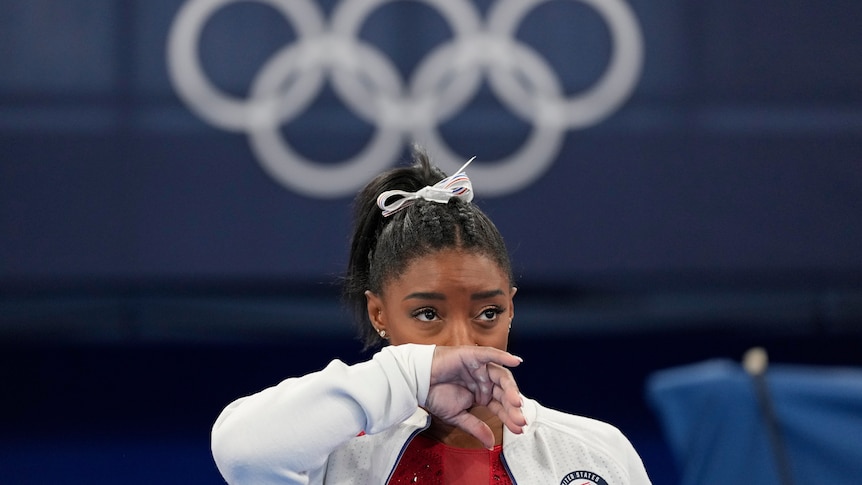 A young woman in a white hoodie wipes her nose. The olympic rings hang on the wall behind her