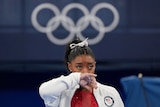 A young woman in a white hoodie wipes her nose. The olympic rings hang on the wall behind her
