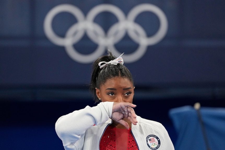 A young woman in a white hoodie wipes her nose. The olympic rings hang on the wall behind her