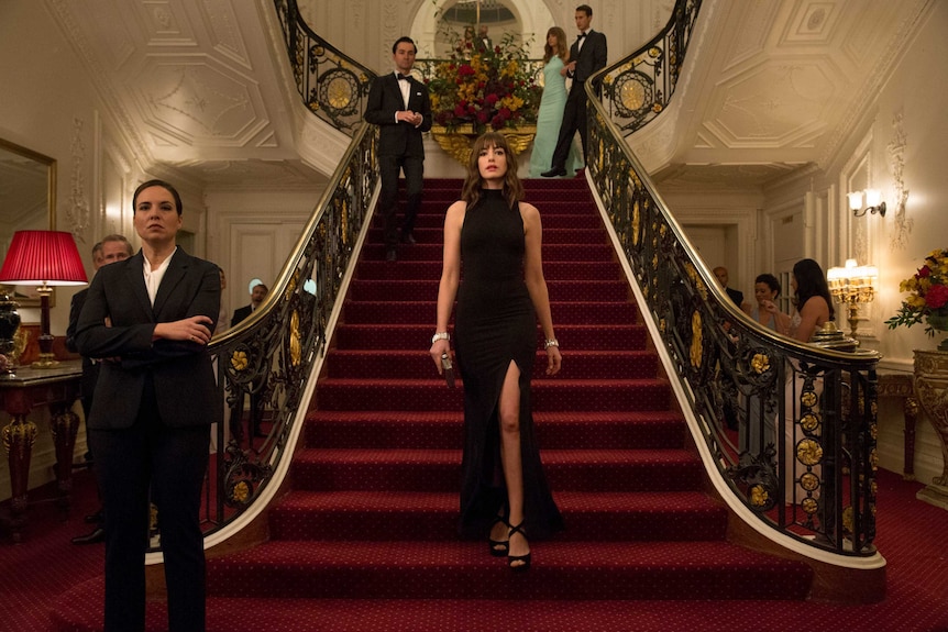 Hathaway stands at the base of a grand staircase wearing an elegant black gown.