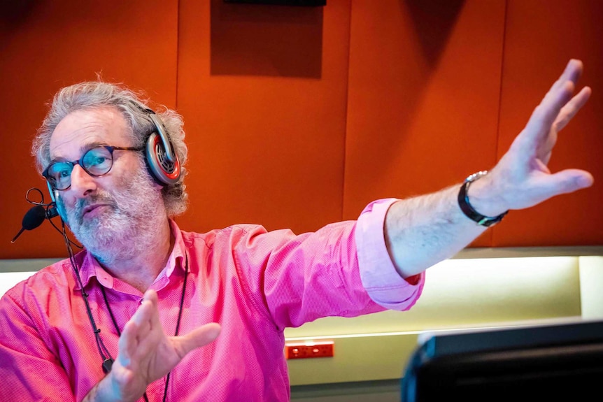 Radio host Jon Faine gestures in from inside a studio with red fabric panels on the walls.