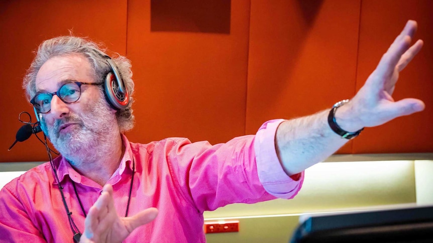 Radio host Jon Faine gestures in from inside a studio with red fabric panels on the walls.