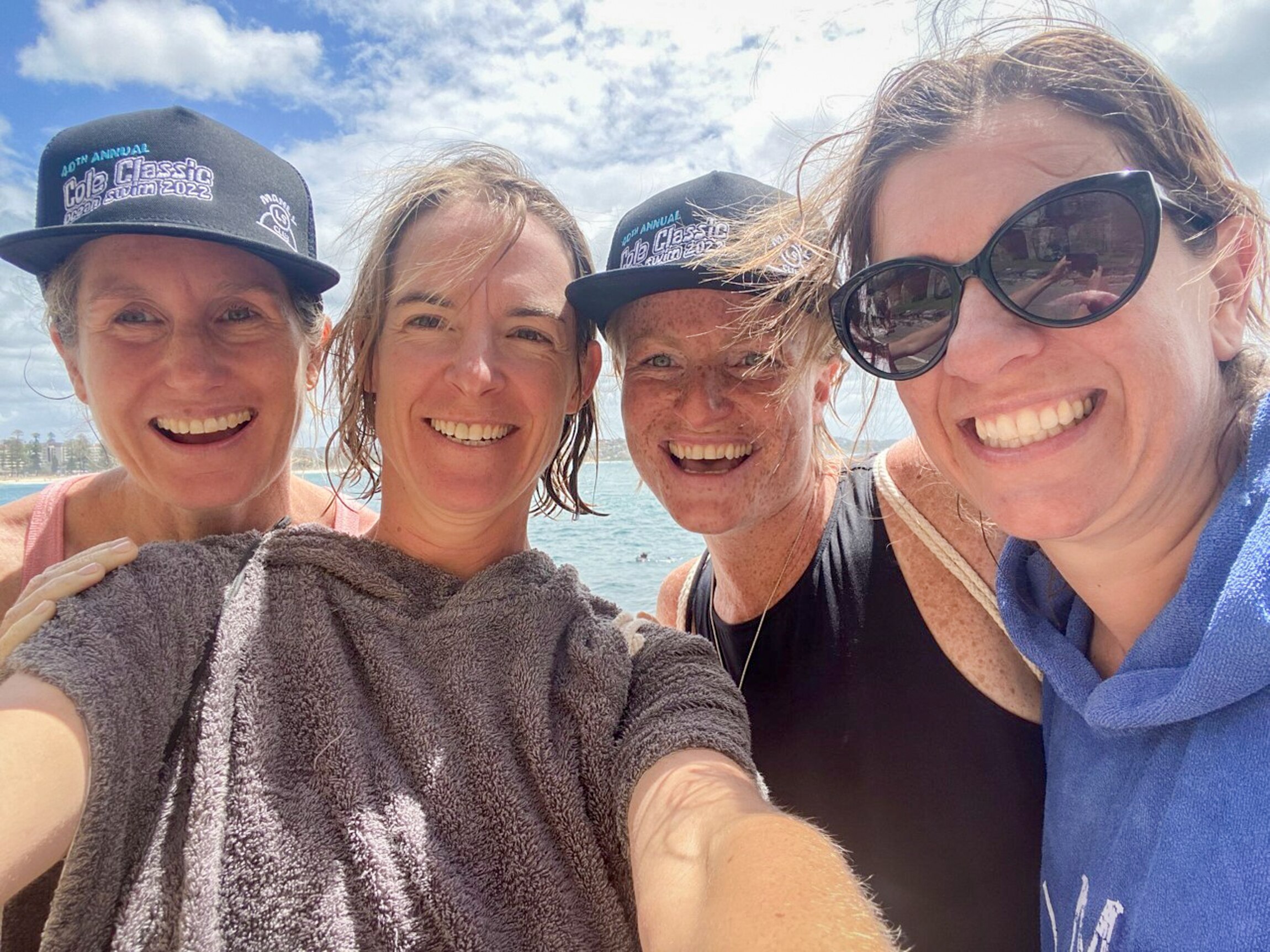 Four women wearing bathers and towels smile at the camera with the blue ocean behind them 