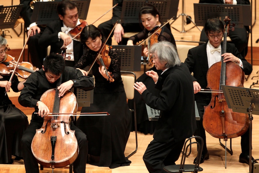 A man wearing a black suit conducting an orchestra where people are holding brown cellos