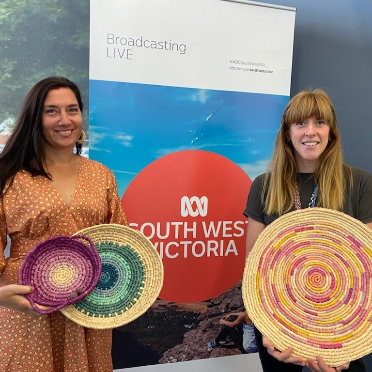 Two women stand holding traditionally woven baskets next to abc south west victoria sign