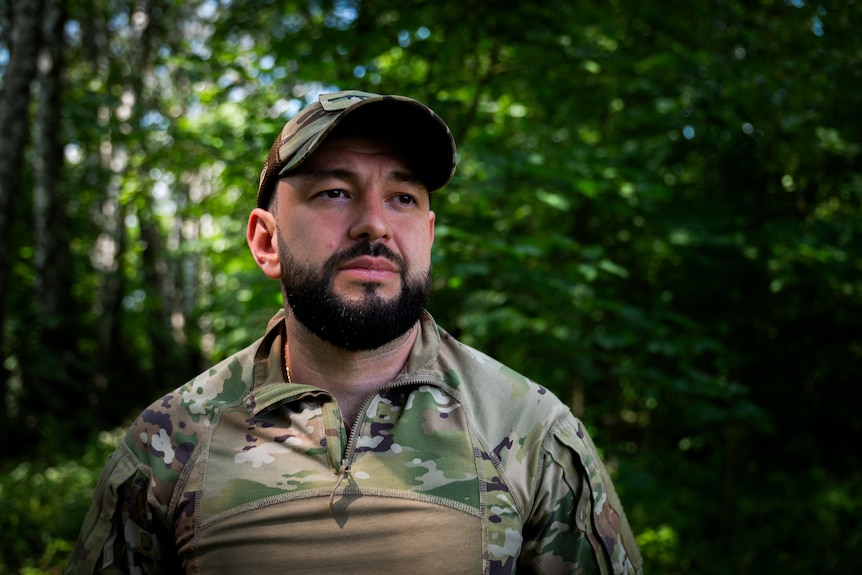 A young man with dark beard, wearing camouflage army fatigues and hat, stands in a forest with green canopy behind him