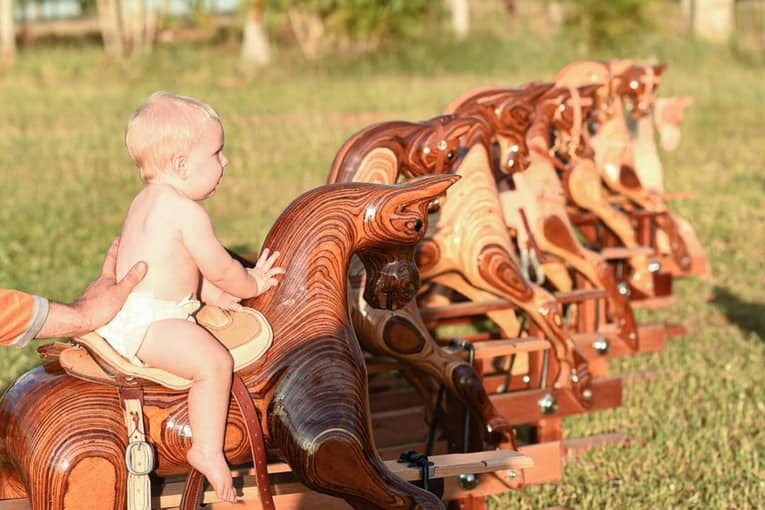 A shirtless, blonde-haired baby in a nappy rides a rocking horse, with a line of other rocking horses behind it.