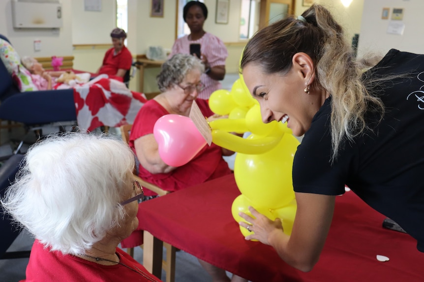 An elderly woman with grey hair looking at a big yellow balloon with a younger woman smiling at her