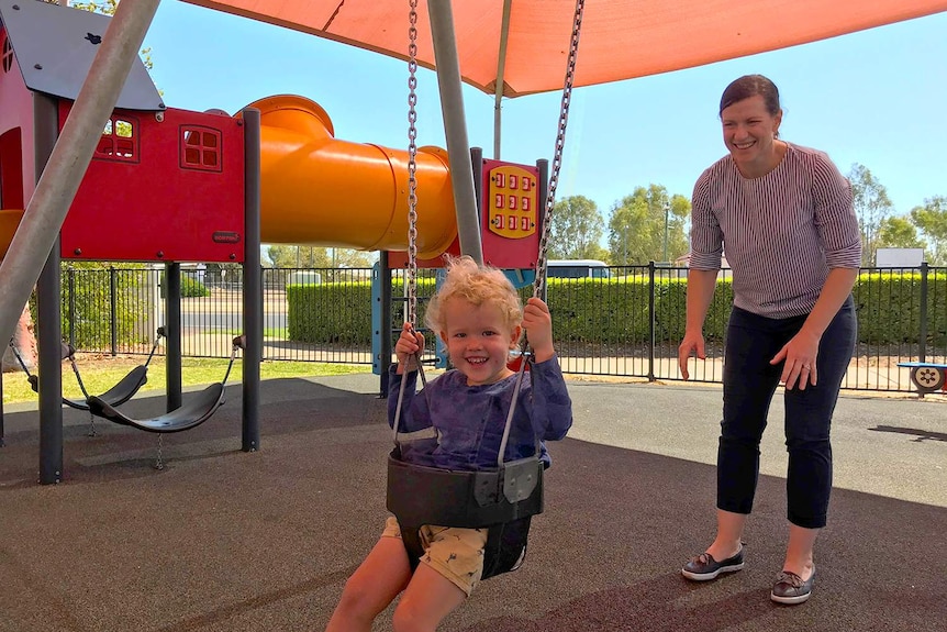 A lady pushes her son on a swing in front of a playground