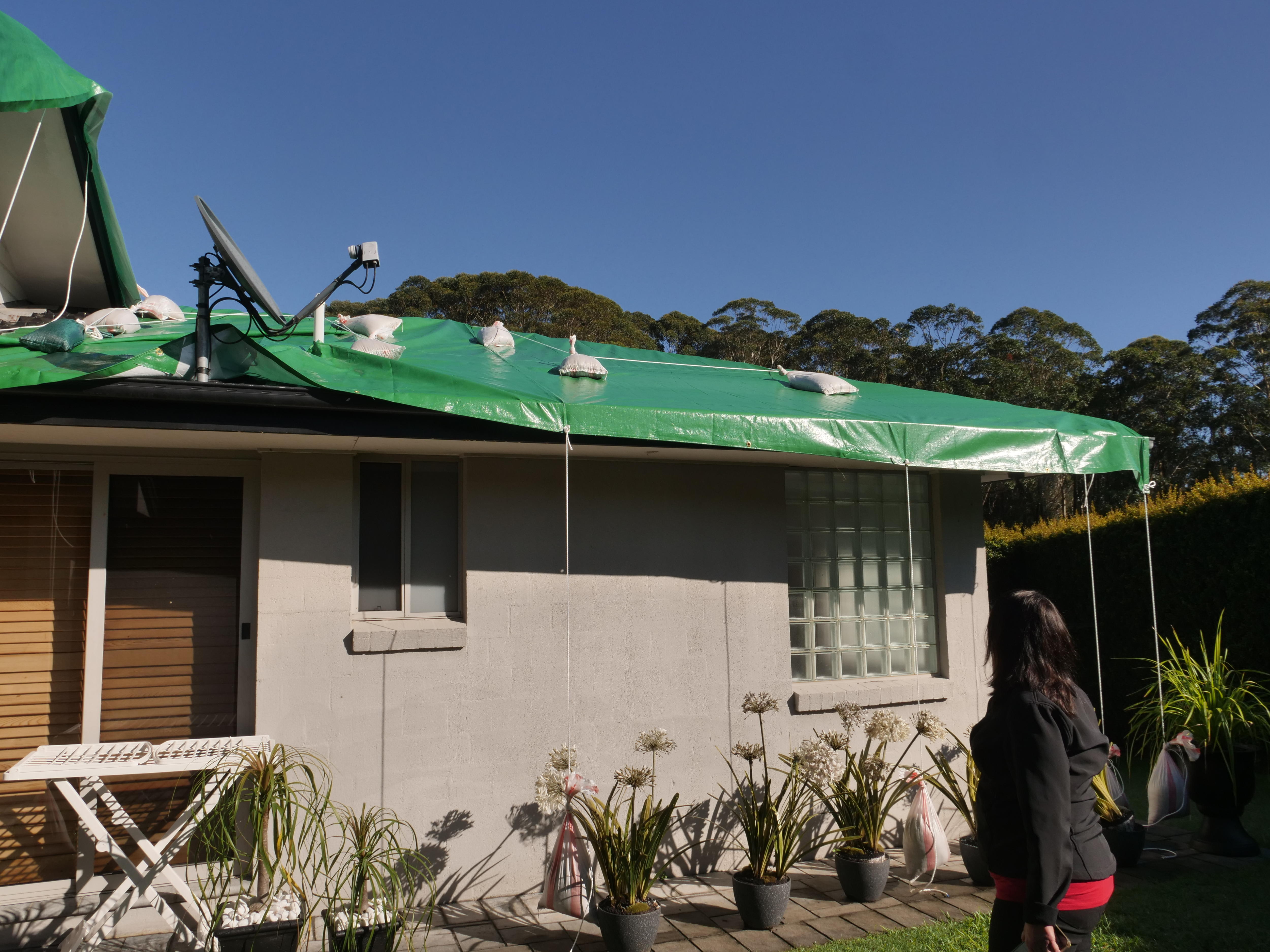 A woman looks at her damaged roof which is covered with a green tarp.