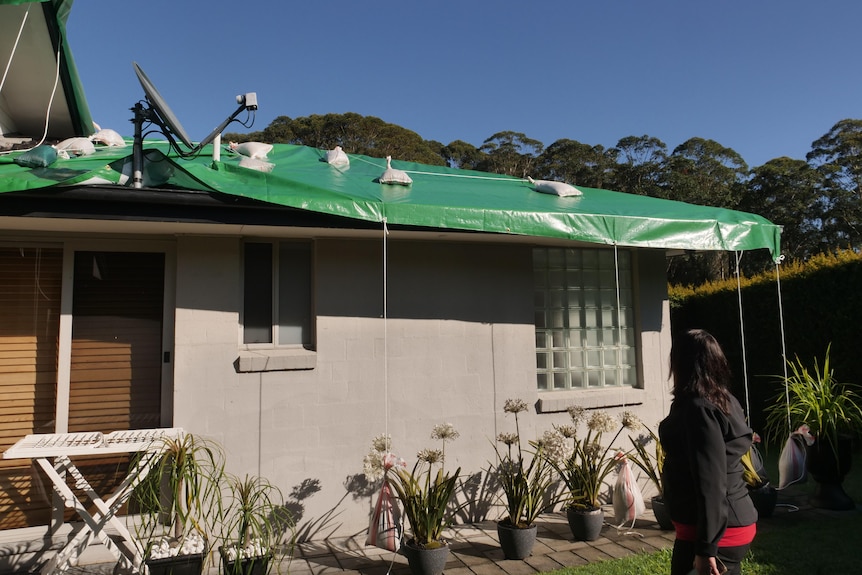 A woman looks at her damaged roof which is covered with a green tarp.