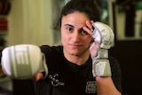 A woman with dark hair, wearing boxing gloves with one hand near her face and the other straight out in front.