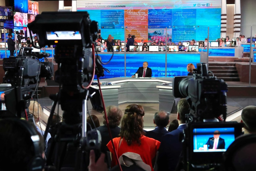 Vladimir Putin sits at a desk in a studio, surrounded by TV cameras and screens.