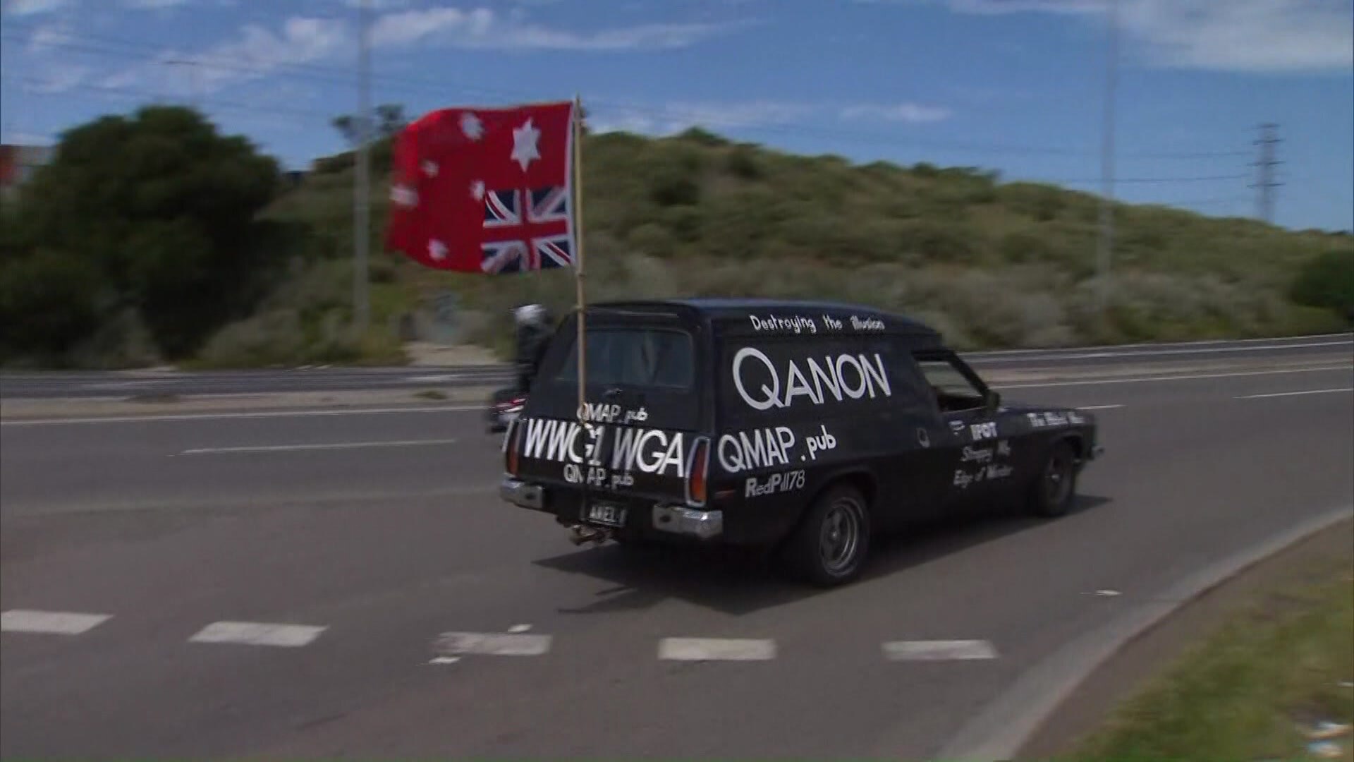 A black car painted in white words relating to QAnon, with an upside down Australian flag flying from the back.