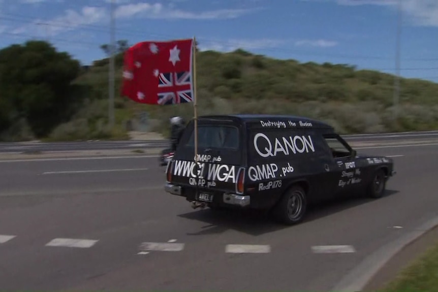 A black car painted in white words relating to QAnon, with an upside down Australian flag flying from the back.