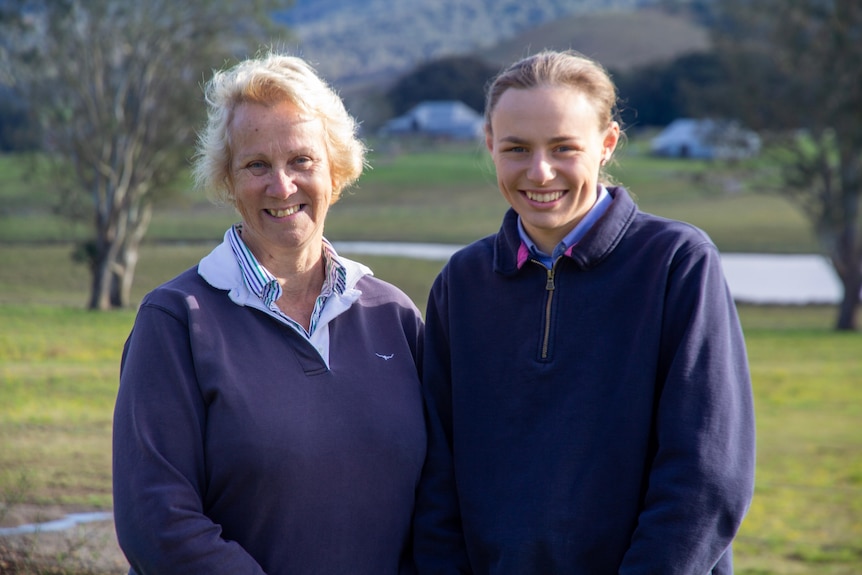 Two women, a grandmother and granddaughter stand together and smile at the camera, standing in front of a farm