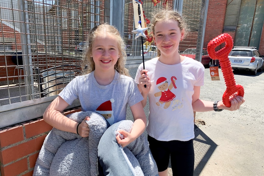 Two young girls stand outside in the sun in a car park holding up two grey cat beds and a wand with a silver star