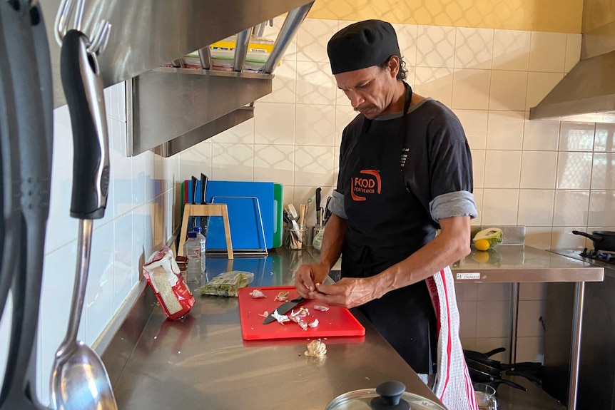 A man in chef hat and apron chops garlic on a chopping board.