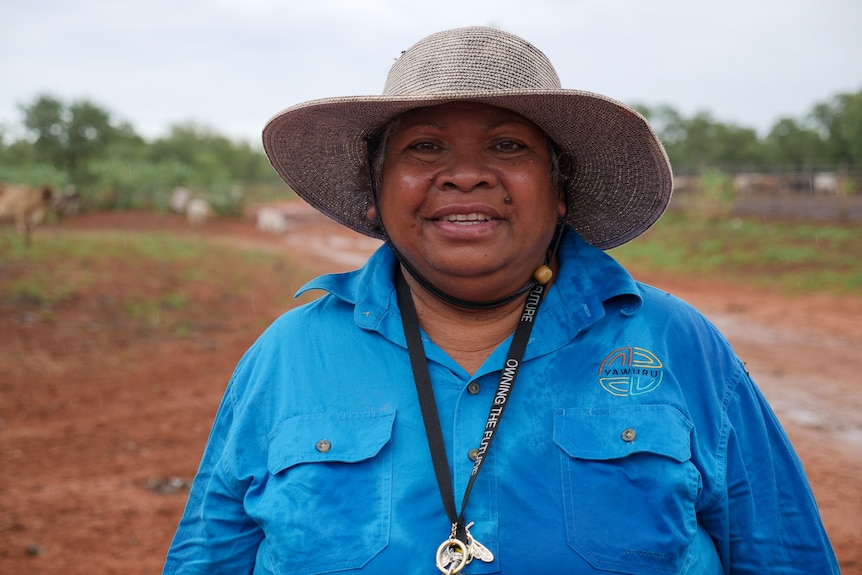 An indigenous woman wearing a cowboy hat and blue shirt standing on red pindan dirt