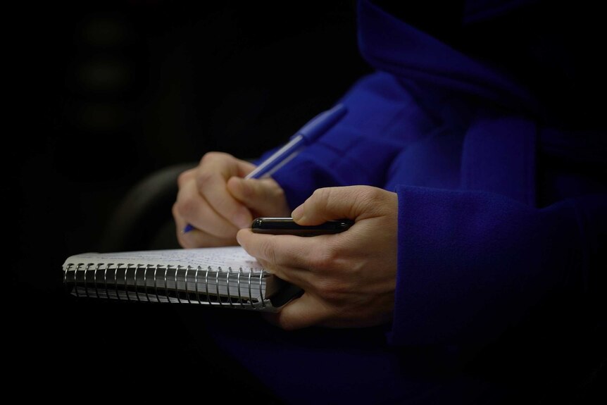Close up of reporter's hands holding phone and writing notes in notebook.