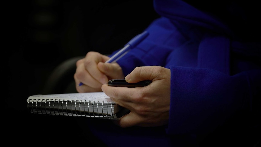 Close up of reporter's hands holding phone and writing notes in notebook.