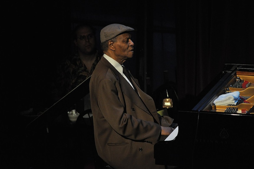 A man wearing a cap and a brown suit sits at a piano.