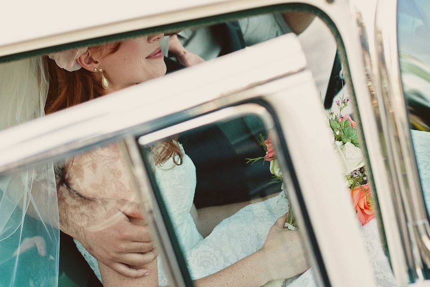 A woman dressed in a wedding dress and veil smiles as she holds as bouquet of flowers in the back seat of a car.